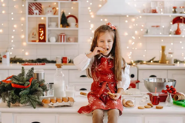 圣诞厨房里一个快乐的小女孩 手里拿着饼干坐在桌子上吃着 — 图库照片