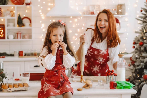 新年厨房里 一个小女孩手里拿着饼干坐在桌子上 妈妈在准备面团 — 图库照片