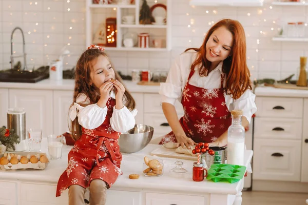 新年厨房里 一个小女孩坐在桌子上 手里拿着饼干吃着 妈妈正在准备面团 — 图库照片