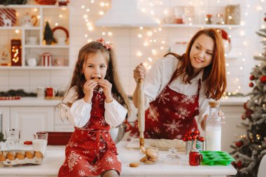 Yeni yıl mutfağında küçük bir kız elinde kurabiyelerle masada oturuyor ve annesi hamur hazırlıyor..