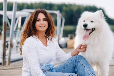 Büyük beyaz bir köpeği olan mutlu bir kadın gün batımında deniz kenarında bir rıhtımda oturur..