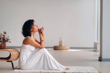 Parlak iç çamaşırlı beyaz elbiseli bir kadın beyaz bir halıda oturuyor. Rahatlama konsepti.