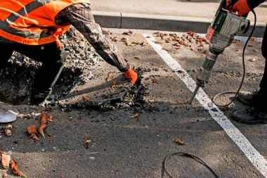 İşçiler sonbahar günü eski bir asfalt tabakasını kaya matkabıyla çıkarırlar. Yolda inşaat işi var..