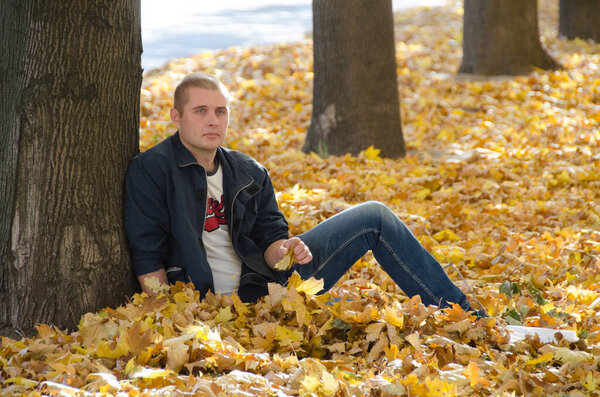 Portrait of a confident man in a black jacket against the backdrop of an autumn park. A man sits on a carpet of yellow autumn leaves.