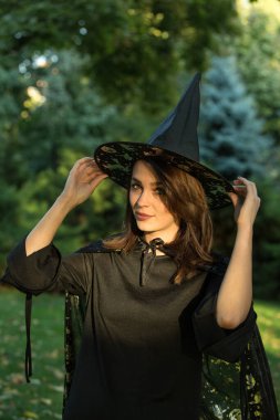 Portrait of attractive young woman in green forest. The girl is wearing a wide-brimmed black pointed hat. A girl with dark hair and hazel green eyes.