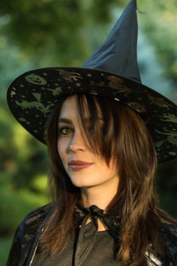 Portrait of attractive young woman in green forest. The girl is wearing a wide-brimmed black pointed hat. A girl with dark hair and hazel green eyes.