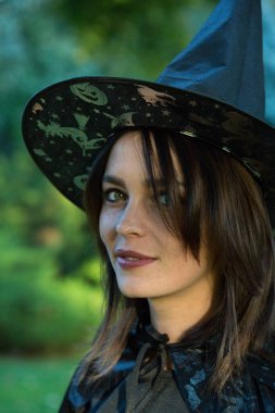 Portrait of attractive young woman in green forest. The girl is wearing a wide-brimmed black pointed hat. A girl with dark hair and hazel green eyes.