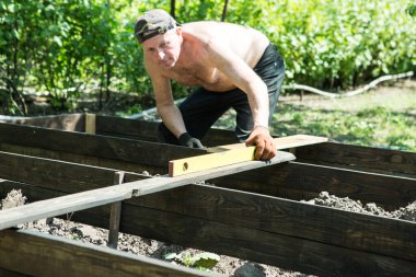 Yetişkin bir adam, kişisel bir komploda yüksek yataklar yapar. Usta, tahtaların doğru boyutlarını korumak için bir seviye aracı ile çalışır.