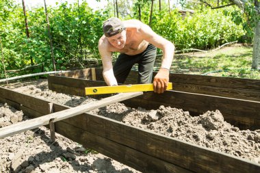 Yetişkin bir adam, kişisel bir komploda yüksek yataklar yapar. Usta, tahtaların doğru boyutlarını korumak için bir seviye aracı ile çalışır.