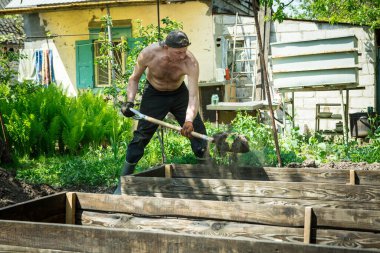 Yetişkin bir adam, kişisel bir komploda yüksek yataklar yapar. Efendi, yatağın ahşap çerçevesine kürekle bereketli toprak döker..