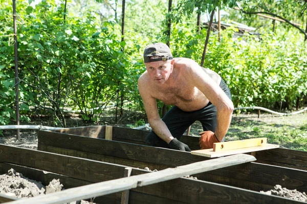 Yetişkin bir adam, kişisel bir komploda yüksek yataklar yapar. Usta, tahtaların doğru boyutlarını korumak için bir seviye aracı ile çalışır.