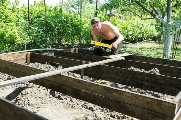 Yetişkin bir adam, kişisel bir komploda yüksek yataklar yapar. Usta, tahtaların doğru boyutlarını korumak için bir seviye aracı ile çalışır.