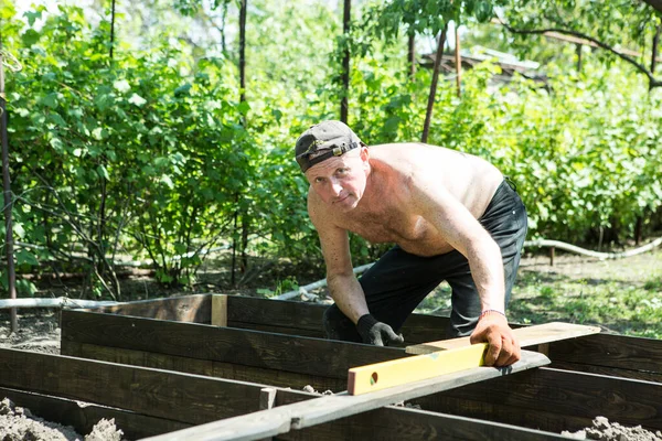 Yetişkin bir adam, kişisel bir komploda yüksek yataklar yapar. Usta, tahtaların doğru boyutlarını korumak için bir seviye aracı ile çalışır.