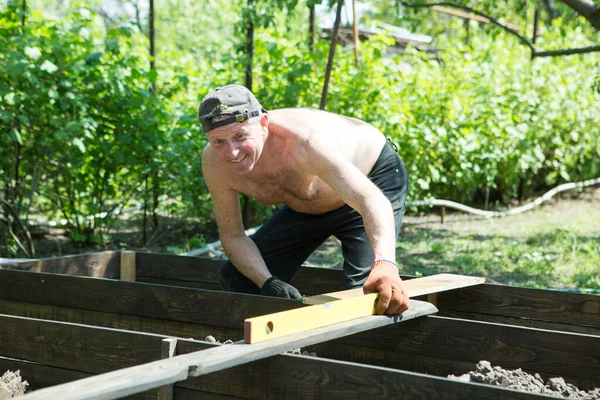 Yetişkin bir adam, kişisel bir komploda yüksek yataklar yapar. Usta, tahtaların doğru boyutlarını korumak için bir seviye aracı ile çalışır.