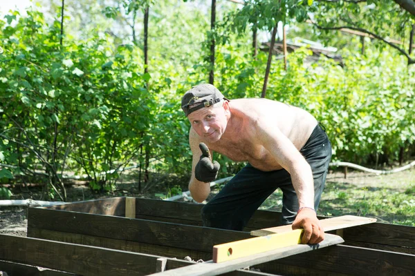 Yetişkin bir adam, kişisel bir komploda yüksek yataklar yapar. Usta, tahtaların doğru boyutlarını korumak için bir seviye aracı ile çalışır.