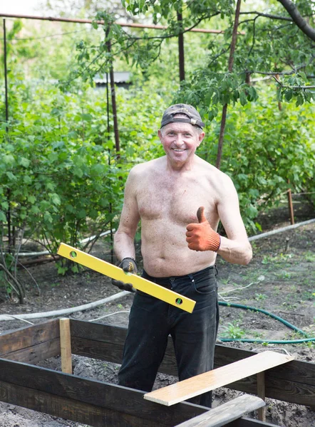 Yetişkin bir adam, kişisel bir komploda yüksek yataklar yapar. Usta, tahtaların doğru boyutlarını korumak için bir seviye aracı ile çalışır.