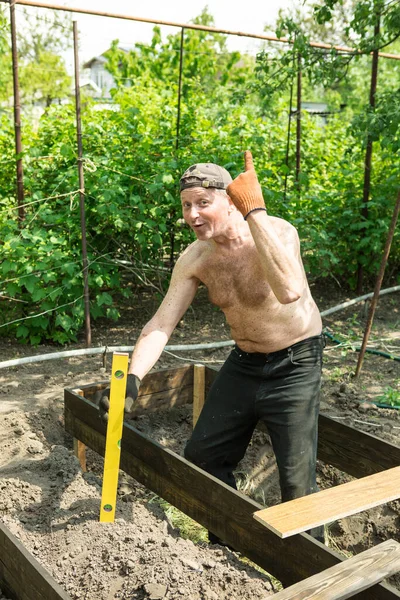 Yetişkin bir adam, kişisel bir komploda yüksek yataklar yapar. Usta, tahtaların doğru boyutlarını korumak için bir seviye aracı ile çalışır.