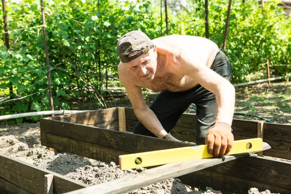 Yetişkin bir adam, kişisel bir komploda yüksek yataklar yapar. Usta, tahtaların doğru boyutlarını korumak için bir seviye aracı ile çalışır.