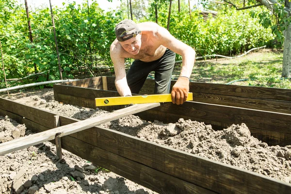 Yetişkin bir adam, kişisel bir komploda yüksek yataklar yapar. Usta, tahtaların doğru boyutlarını korumak için bir seviye aracı ile çalışır.