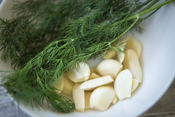 One bunch of fresh green dill and peeled garlic cloves on a white plate close-up. Selective focus.