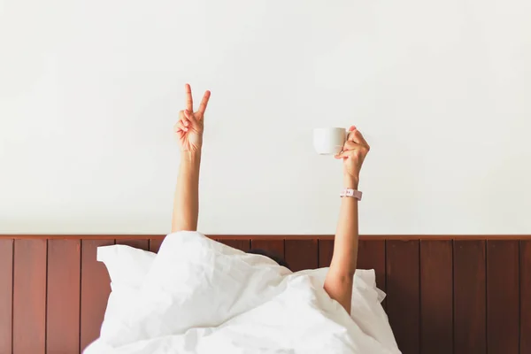 Mano femminile mostrando il segno divertente e tenendo la tazza di caffè dietro la coperta nel letto. — Foto Stock