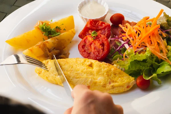 Kvinnlig hand håller gaffel och kniv äter frukost med omelett potatis, tomater och grönsakssallad på bordet. — Stockfoto