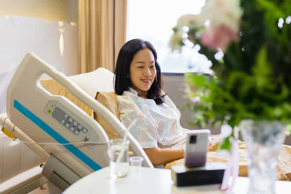 Asiática paciente sonriendo mientras se sienta en la cama haciendo videollamada en el teléfono celular en la sala de hospital. — Foto de Stock