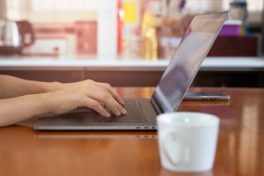 Woman types on the laptop keyboard sitting on table near the window at home.