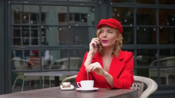 Стильная блондинка в красной кепке и в костюме разговаривает по телефону в кафе на открытом воздухе — стоковое видео