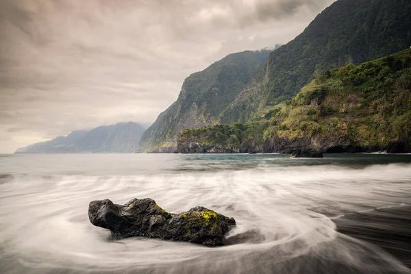 Rock and water lines in a black sand beach with moody cliffs on the background in Seixal (Madeira, Portugal)