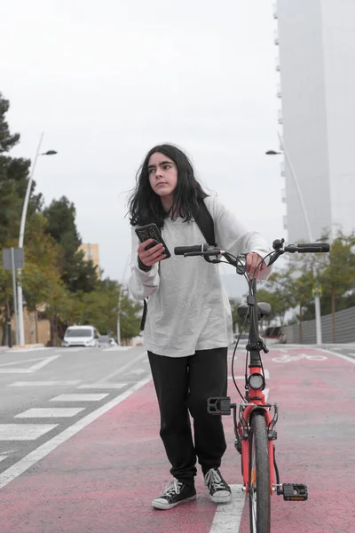 Vertical image, low angle, long shot, of a teenager walking along a bike path pushing a red bicycle while consulting his smart phone. He has long hair and looks to the side.
