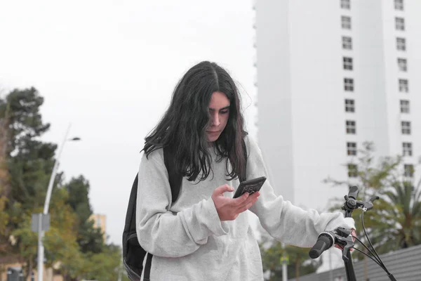 Medium shot horizontal image of a teenager with long hair consulting his cell phone while holding the handlebars of a bicycle, in the background out of focus appears a white skyscraper on a street in a big city.