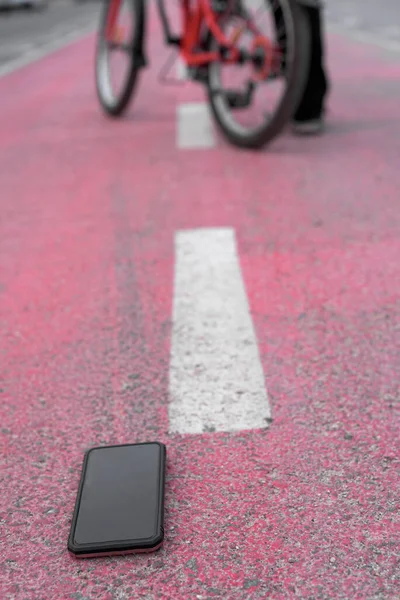 Vertical image, mobile phone is on the floor of a bike path, in the background out of focus there is a bike from which a person comes down to pick up the phone that has fallen.