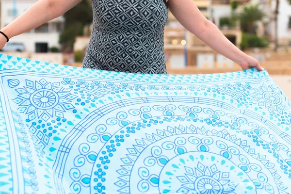 Close-up of a woman in printed dress spreads blue and white towel on the beach, her face is not visible.