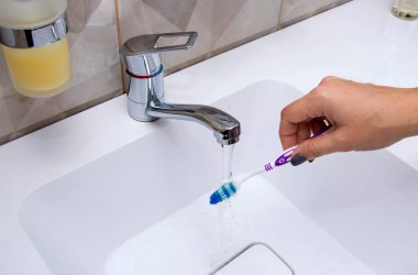 Musluktan akan suyun altında diş fırçası ve diş macunu tutan bir kadın eli.