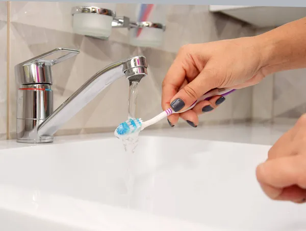 Μια γυναίκα κρατάει μια οδοντόβουρτσα και την πλένει κάτω από το νερό στο νιπτήρα του μπάνιου κάτω από τη βρύση.. — Φωτογραφία Αρχείου