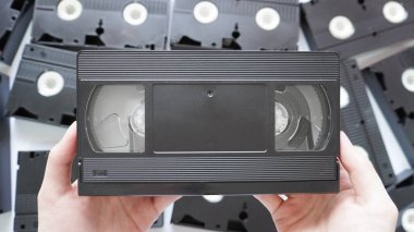 Eller ön tarafta bir VHS kaseti tutuyor. Etrafı dağınık kasetlerle çevrili..