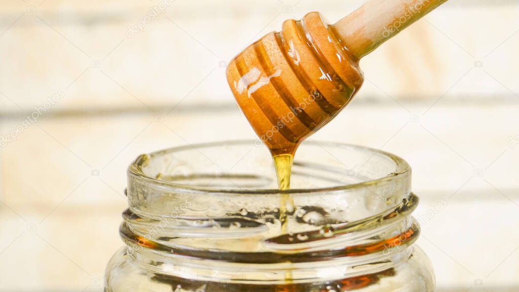 Honey dripping from a honey dipper into a glass honey jar with wood background.