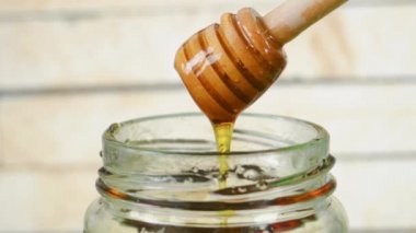 Honey dripping from a honey dipper into a glass honey jar with wood background.