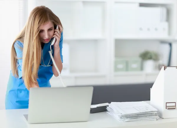 Молодая женщина врач в белом халате за компьютером с помощью телефона — стоковое фото