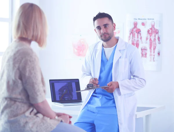 Beau médecin parle avec une jeune patiente et prend des notes alors qu'il est assis dans son bureau. — Photo