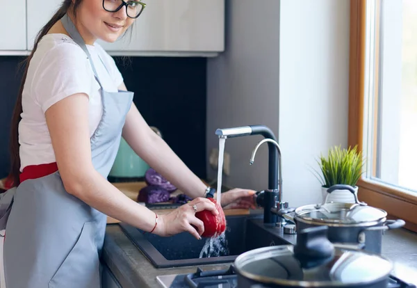 Kvinnan tvättar grönsaker. Vacker ung kvinna som tvättar grönsaker för sallad och ler stående i köket — Stockfoto