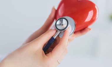 Steteskoplu bir doktor, beyaz arka planda kırmızı kalbi inceliyor.