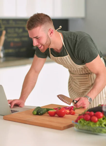 Retrato del hombre cocinando verduras en la cocina mientras mira una computadora portátil en la mesa — Foto de Stock