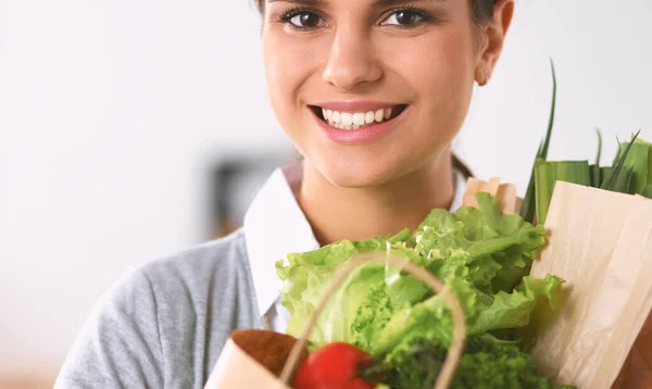 Mujer joven sosteniendo bolsa de la compra de comestibles con verduras de pie en la cocina. —  Fotos de Stock