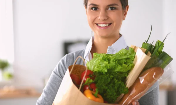 野菜と食料品の買い物袋を持っている若い女性キッチンに立つ. — ストック写真