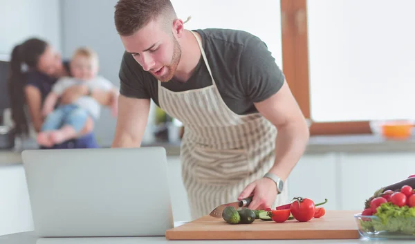 Hombre joven cortando verduras y mujer de pie con el ordenador portátil en la cocina — Foto de Stock