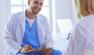 Yakışıklı doktor genç bayan hastasıyla konuşuyor ve ofisinde otururken notlar alıyor.