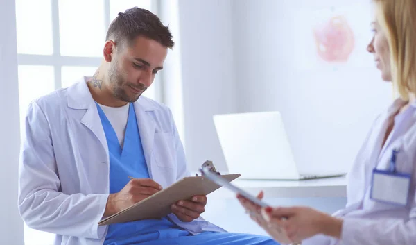Knappe dokter praat met jonge vrouwelijke patiënt en maakt aantekeningen terwijl hij in zijn kantoor zit — Stockfoto