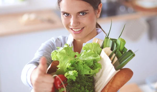 Mujer joven sosteniendo bolsa de la compra de comestibles con verduras. De pie en la cocina — Foto de Stock